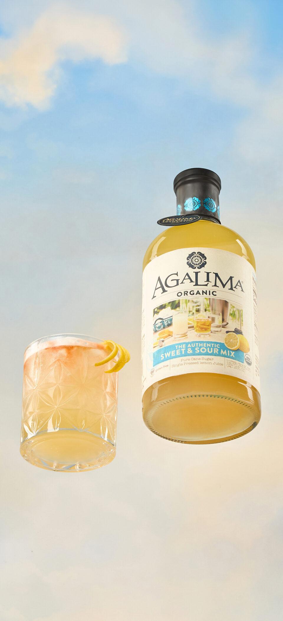 A bottle of Agalima Organic Sweet & Sour Mix floating beside a cocktail garnished with lemon against a soft blue sky
