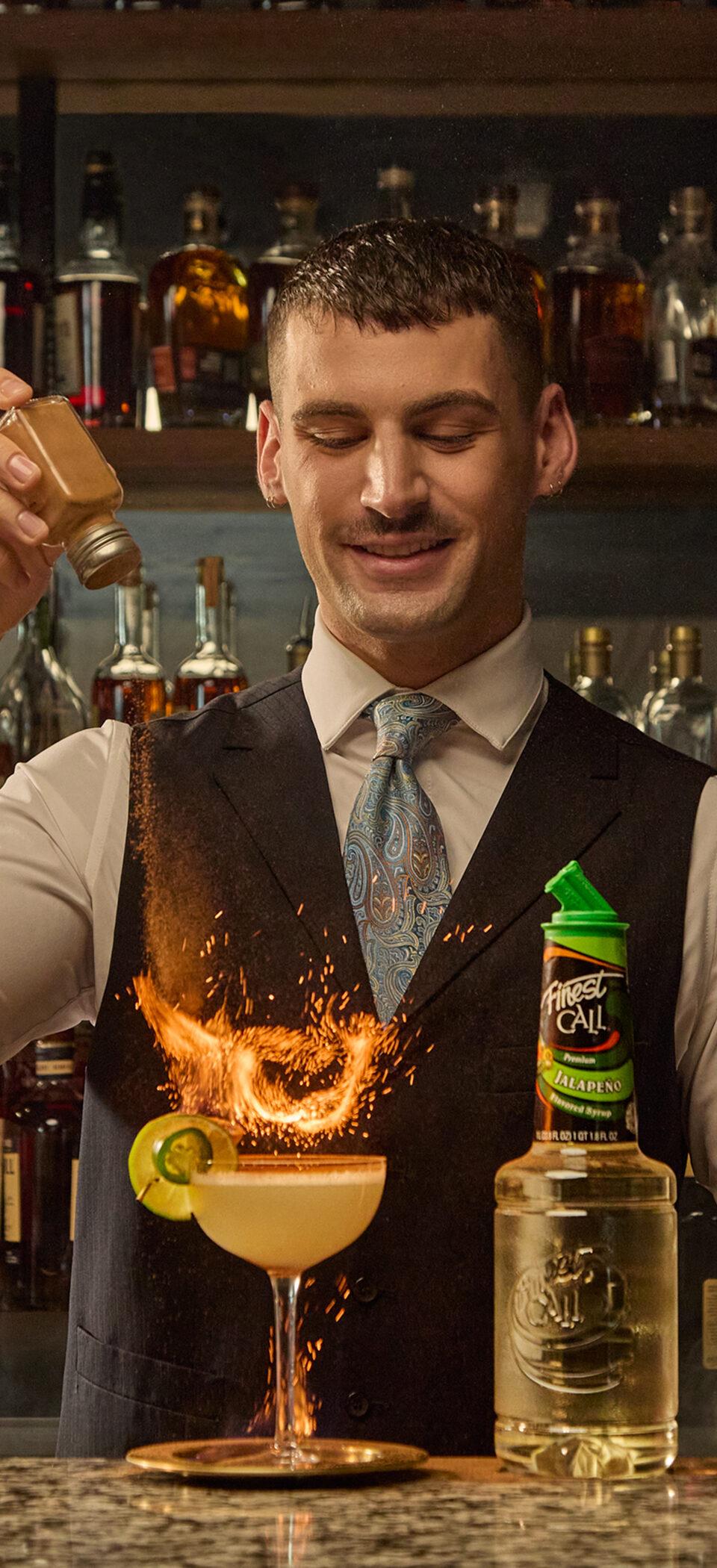 A bartender in a vest and tie sprinkles spice over a flaming cocktail beside a Finest Call Jalapeño mixer at a backlit bar