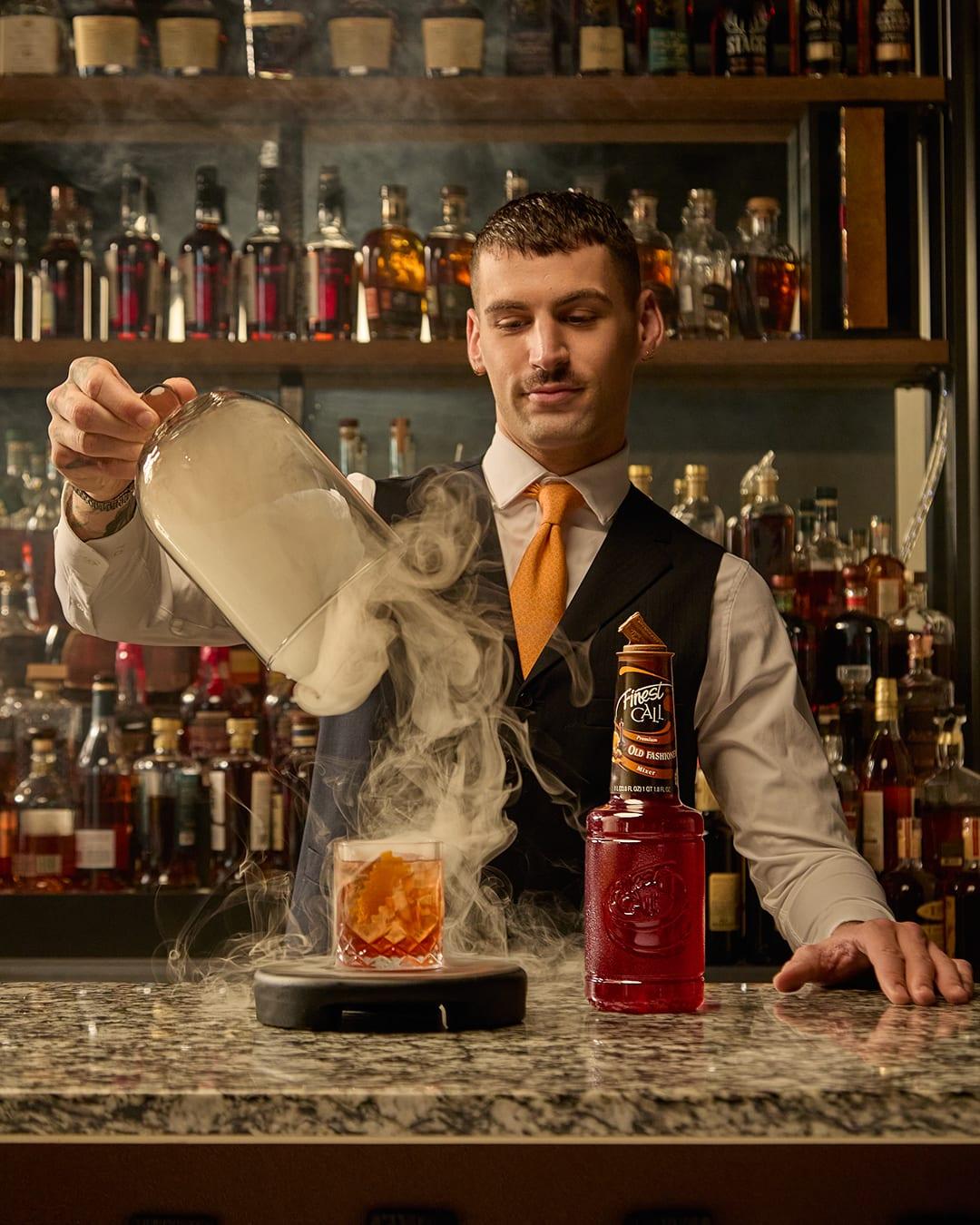A bartender lifts a glass cloche releasing smoke from an old fashioned beside a Finest Call Old Fashioned mixer