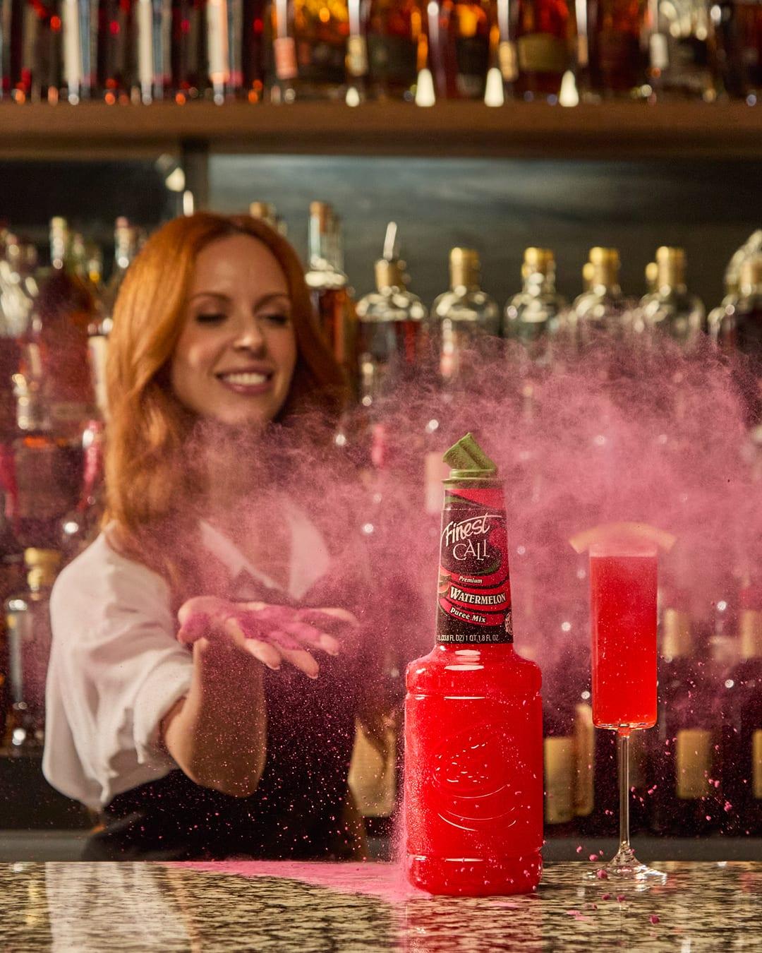 A bartender blows pink powder over a Finest Call Watermelon Purée Mix bottle and a pink cocktail