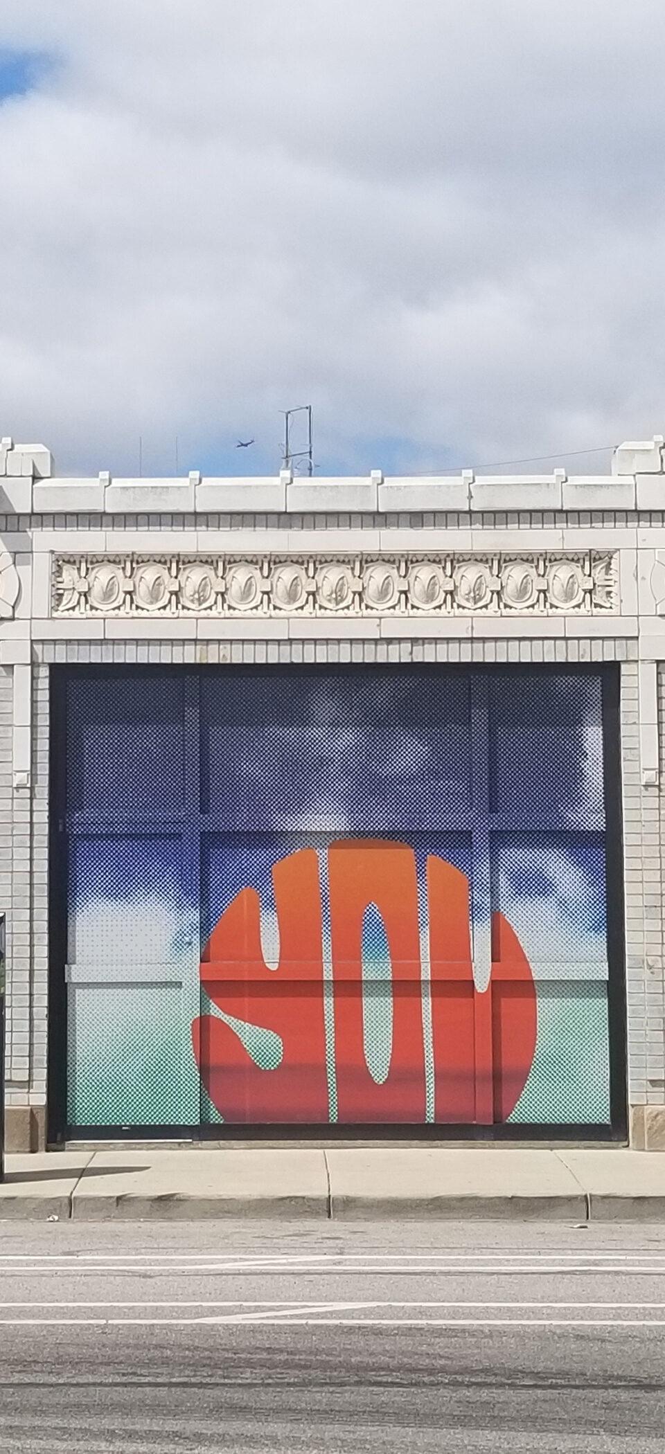 Storefront windows on an Indianapolis street wrapped in IndyArts murals, with bold orange and red lettering over a sky-blue backdrop