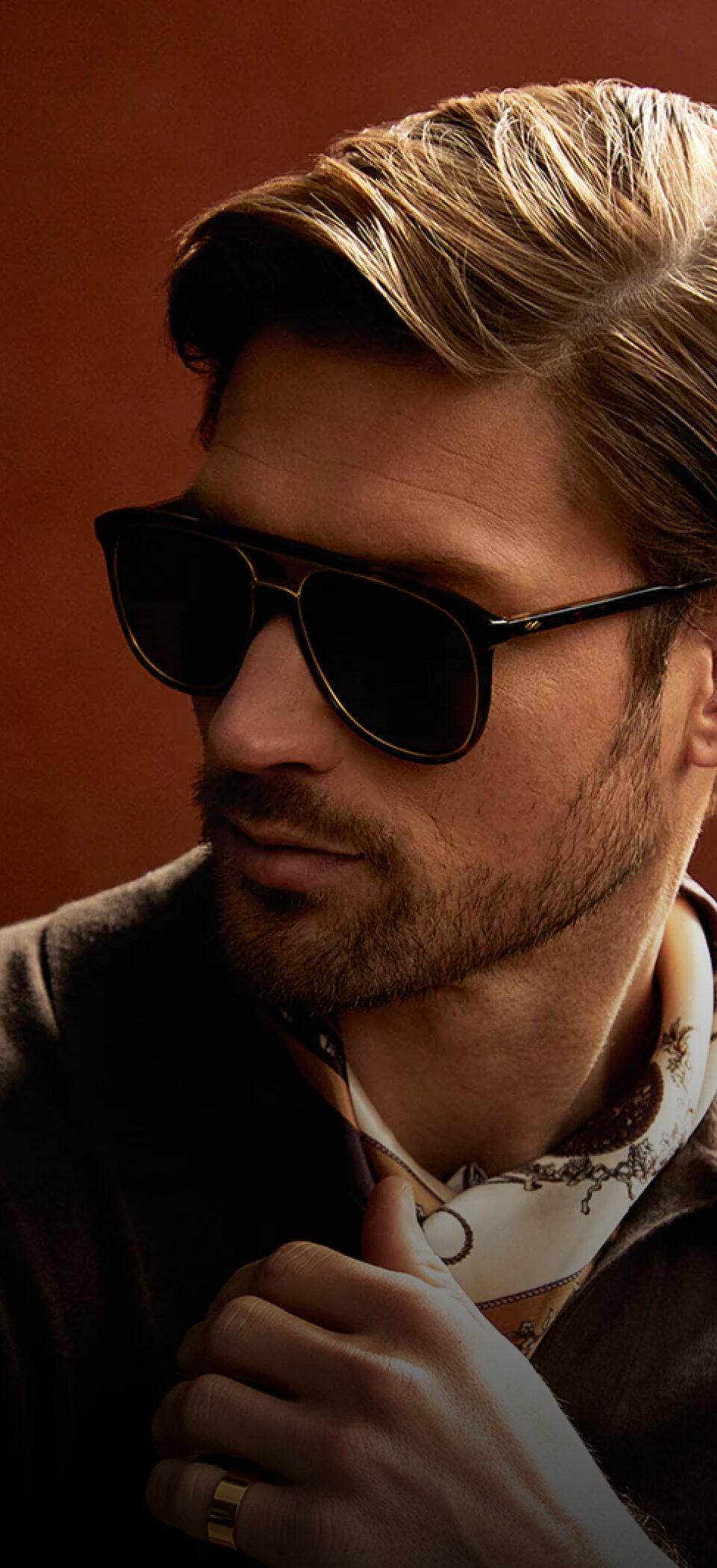 A man with swept-back hair in dark Valdo sunglasses and a patterned neck scarf, set against a deep red backdrop