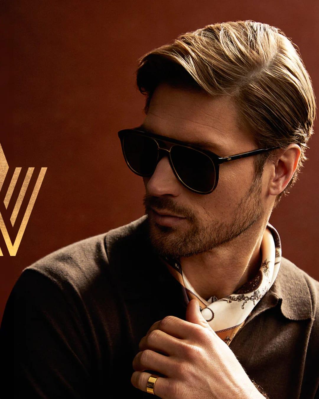 A man in dark Valdo sunglasses and a gold ring beside the bronze Valdo V mark on a red backdrop