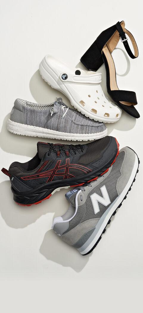 Overhead flat lay of shoes on a pale background: a grey New Balance sneaker, a black and red trail runner, a grey knit slip-on, a white Crocs clog and a black sandal