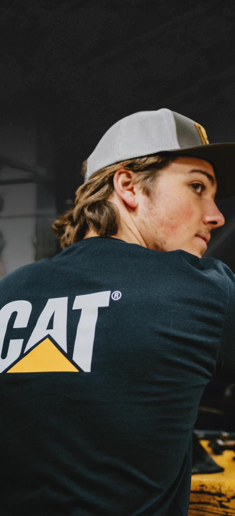 Young worker in a grey cap and black CAT T-shirt looking over his shoulder beside yellow heavy equipment