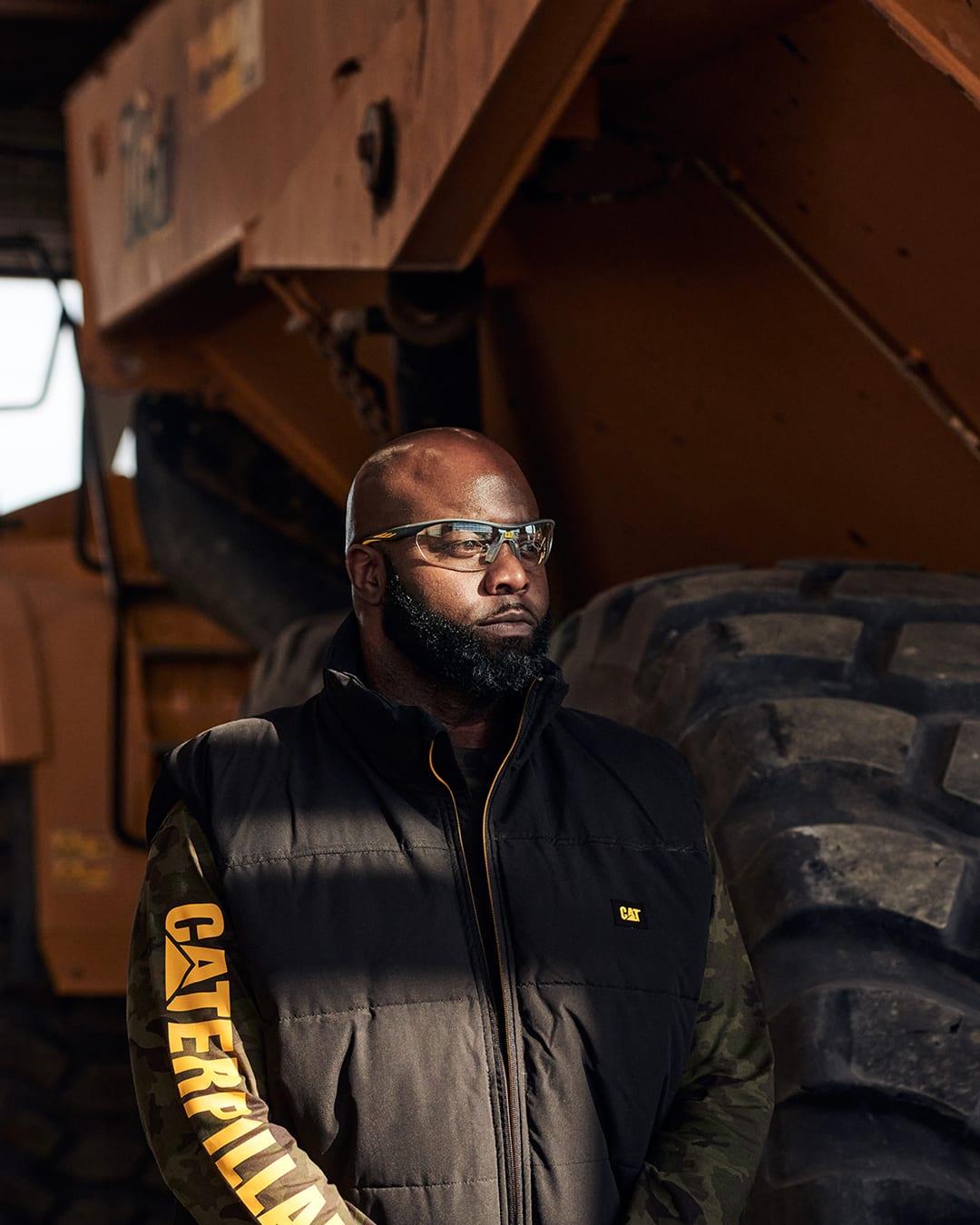Bearded worker in safety glasses and a black CAT vest standing beside a giant earthmover tire