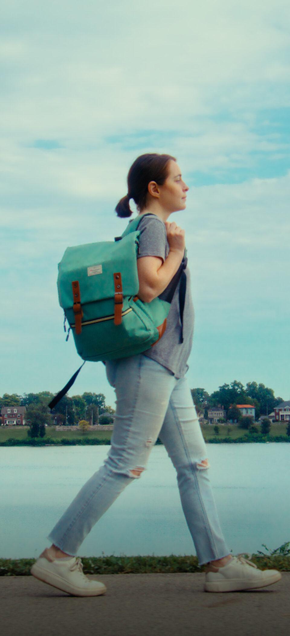 A woman carrying a green backpack walks along a riverside path beneath a bridge in downtown Louisville