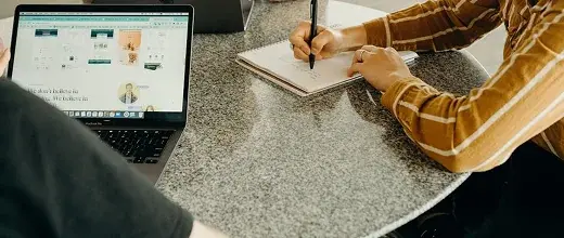 A person taking notes in a notebook beside an open laptop