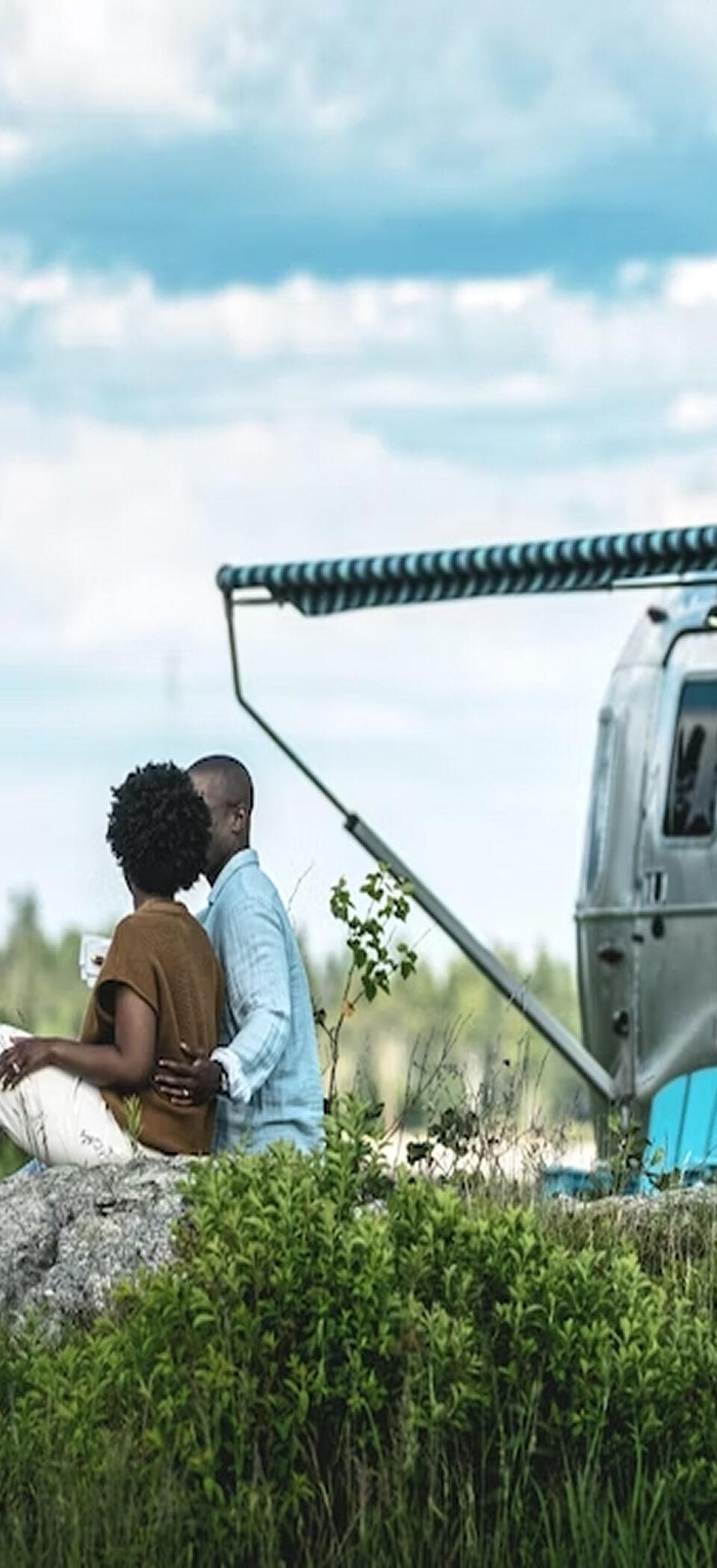 A couple sits on a rock in tall grass beside a silver Airstream travel trailer with its awning extended