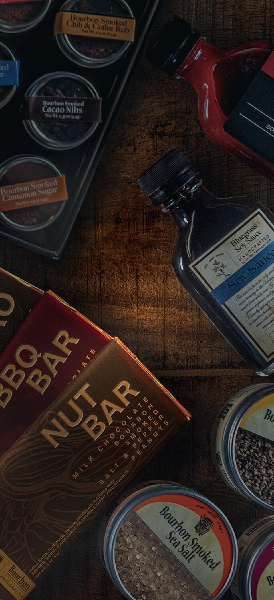 Bourbon Barrel Foods products on a dark wood table: chocolate bars, a bottle of Bluegrass Soy Sauce, finishing sugars and a tin of Bourbon Smoked Pepper