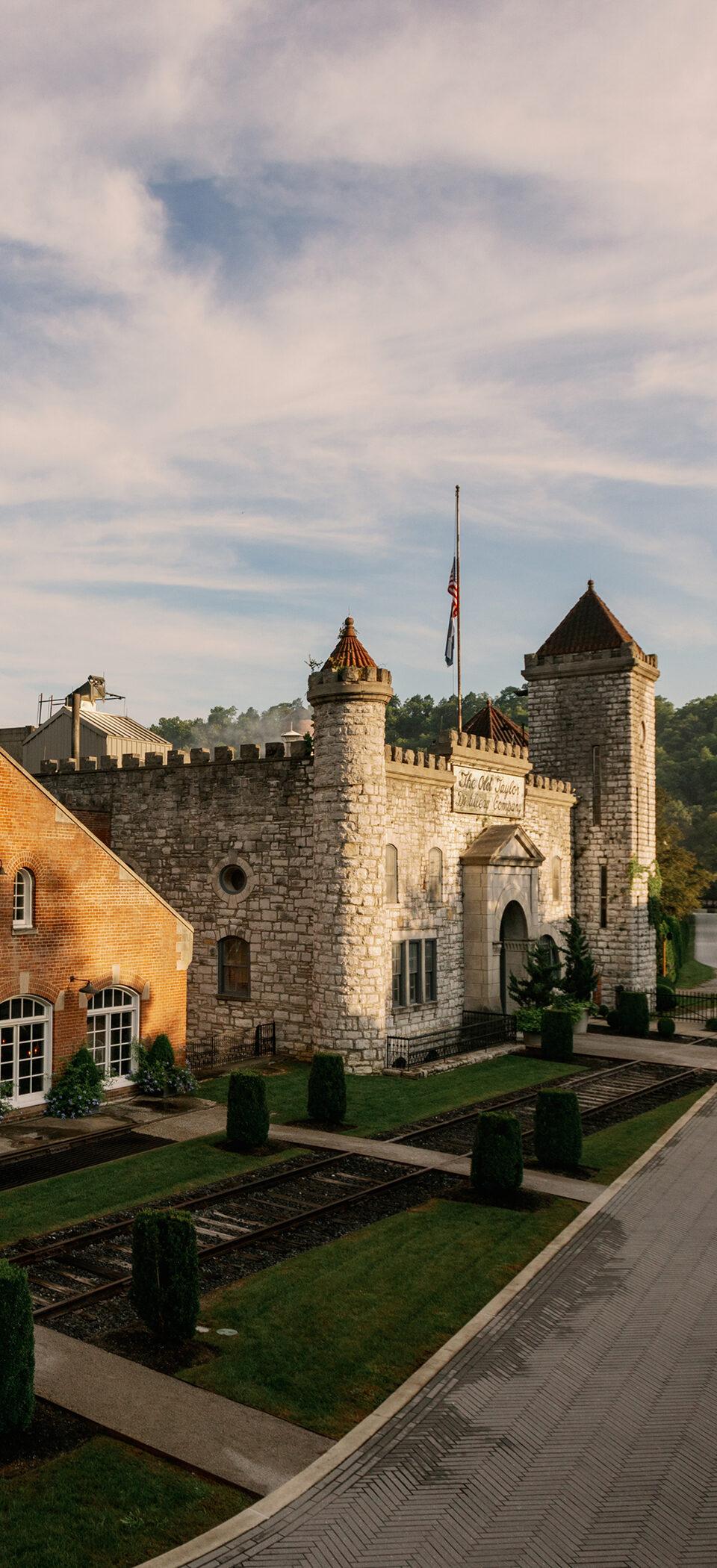 The Castle & Key Distillery grounds at golden hour: a brick smokestack and stone castle buildings beside a manicured garden walk