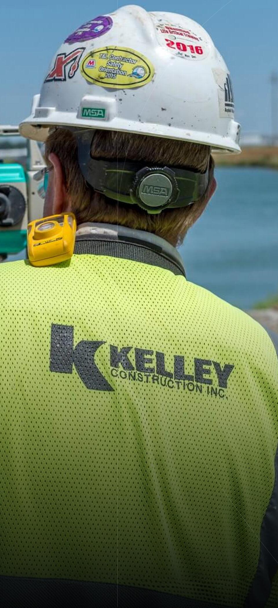 A Kelley Construction worker in a hard hat and high-visibility vest stands at a surveying instrument on a waterfront job site