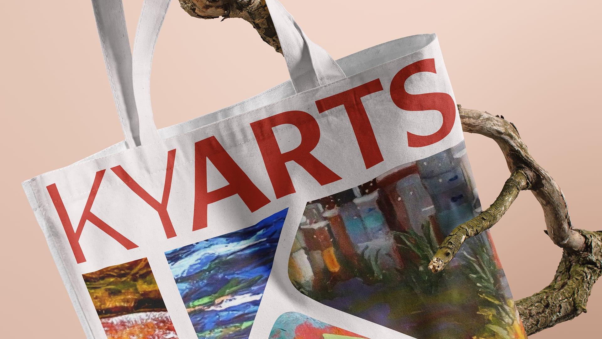 Canvas tote bag printed with the red KYARTS wordmark and artwork, hanging from a branch