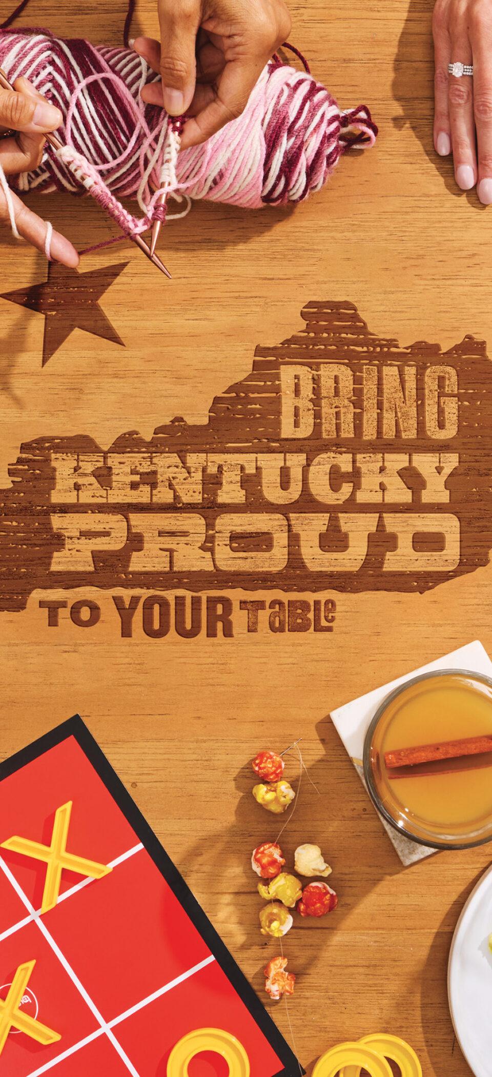 Overhead view of a wooden table with the Kentucky Proud state outline and "Bring Kentucky Proud to your table" burned into the wood, surrounded by hands knitting, playing tic-tac-toe and cards, and plates of pie, fruit, cheese, crackers and cranberry sauce.