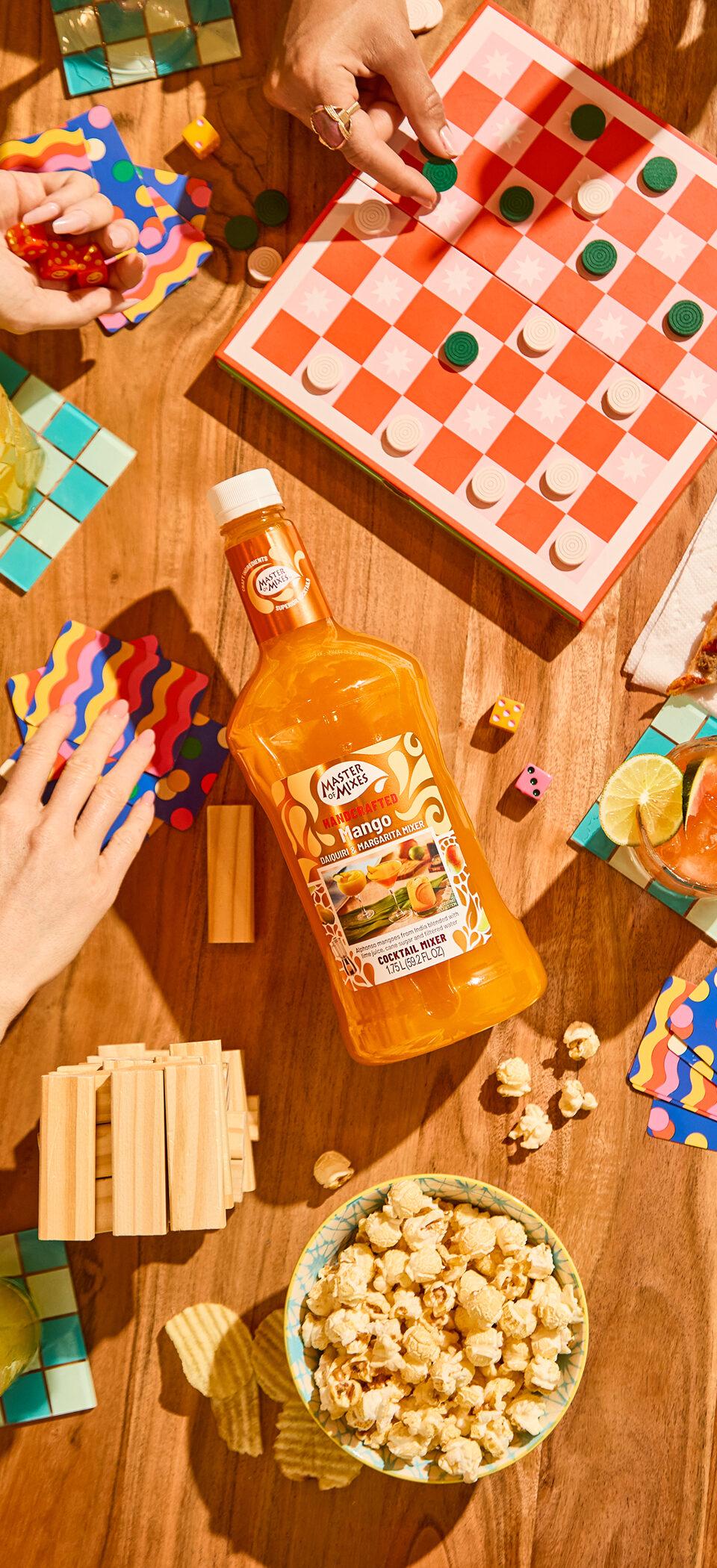 Overhead view of a game night table with a Master of Mixes bottle, popcorn, cocktails and a checkers board