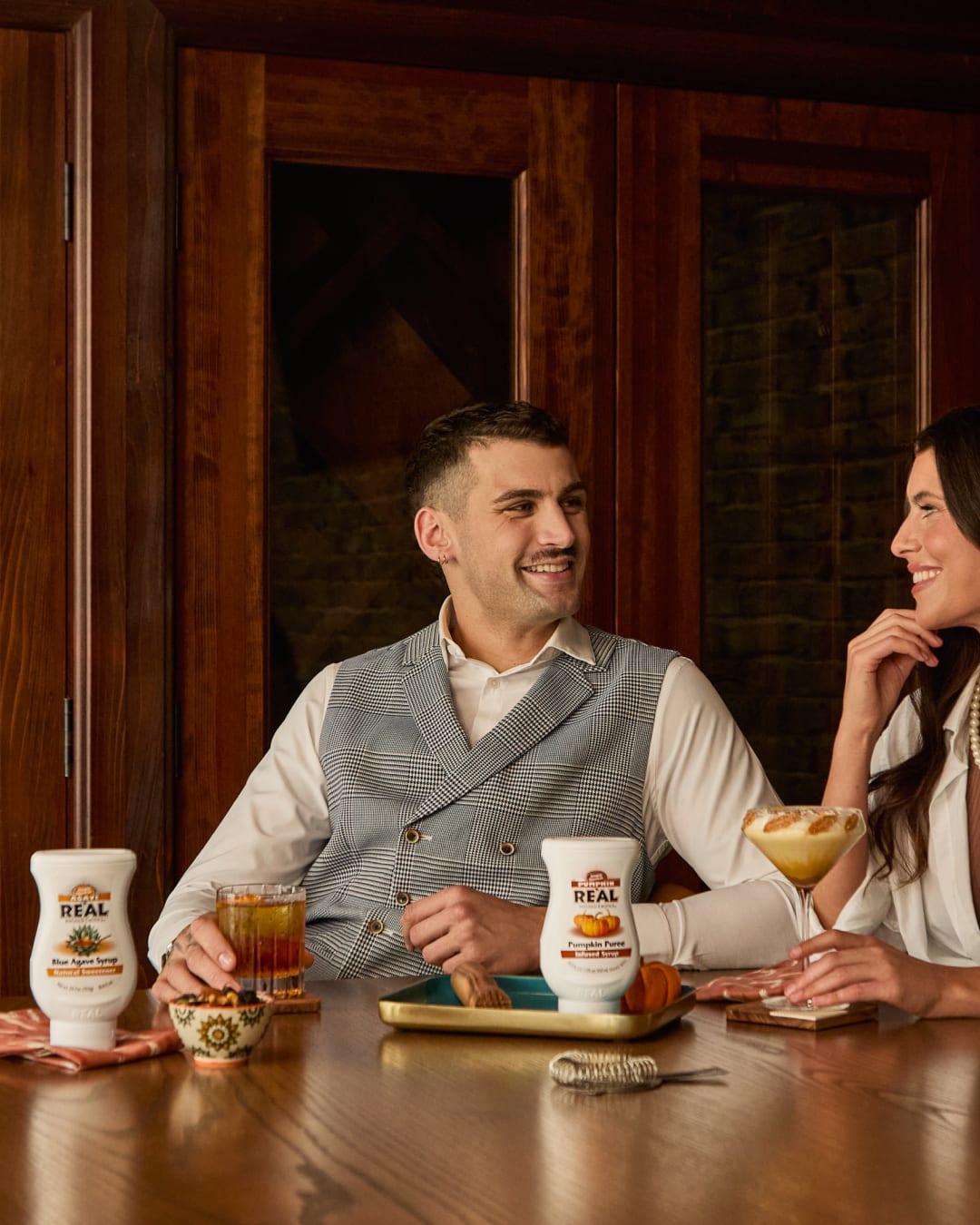 A man and a woman sharing cocktails at a wooden bar with bottles of Reàl syrup